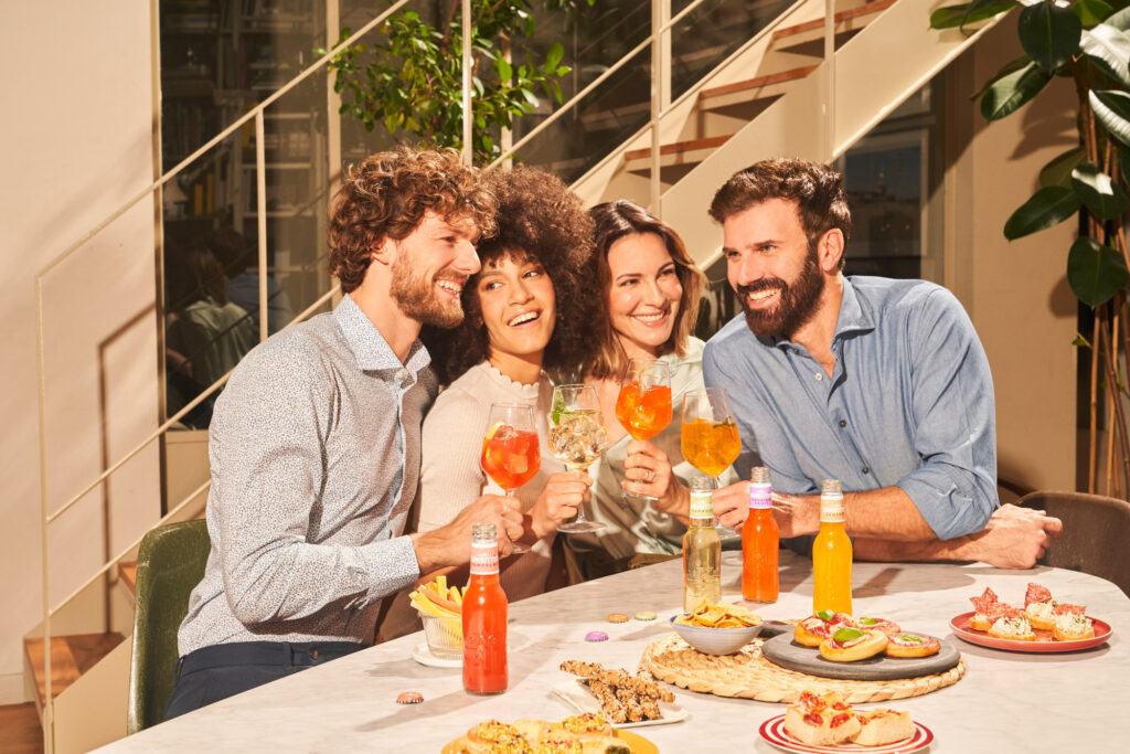 Friends gathered at a beautifully set table, sharing Sanbittèr and an array of snacks. Capturing warmth and connection, ideal for advertisements showcasing the brand’s essence.