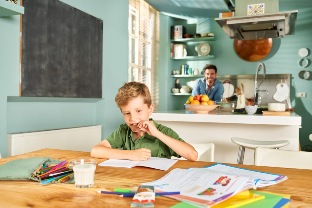 A young boy enjoying a Kinder Cards snack while studying, with a bright kitchen and a smiling father in the background