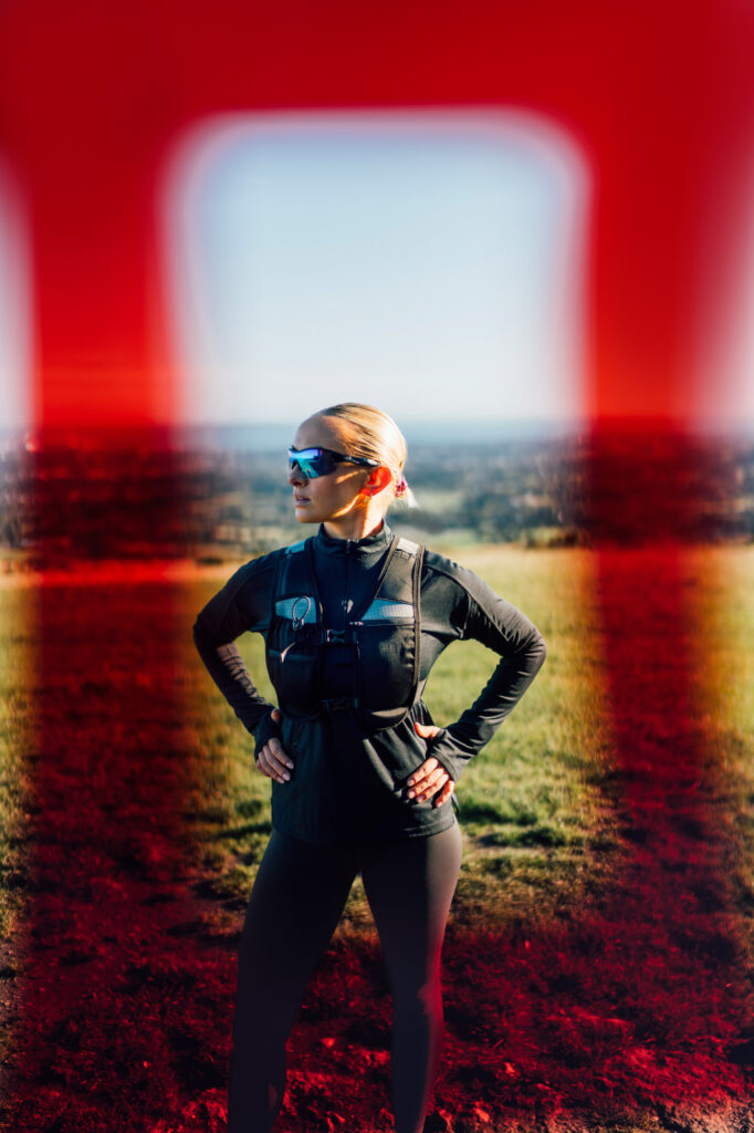 A female trail runner standing in the Irish countryside under a clear blue sky. Her confident posture and the scenic backdrop reflect joy and fitness in nature. Red gel framed