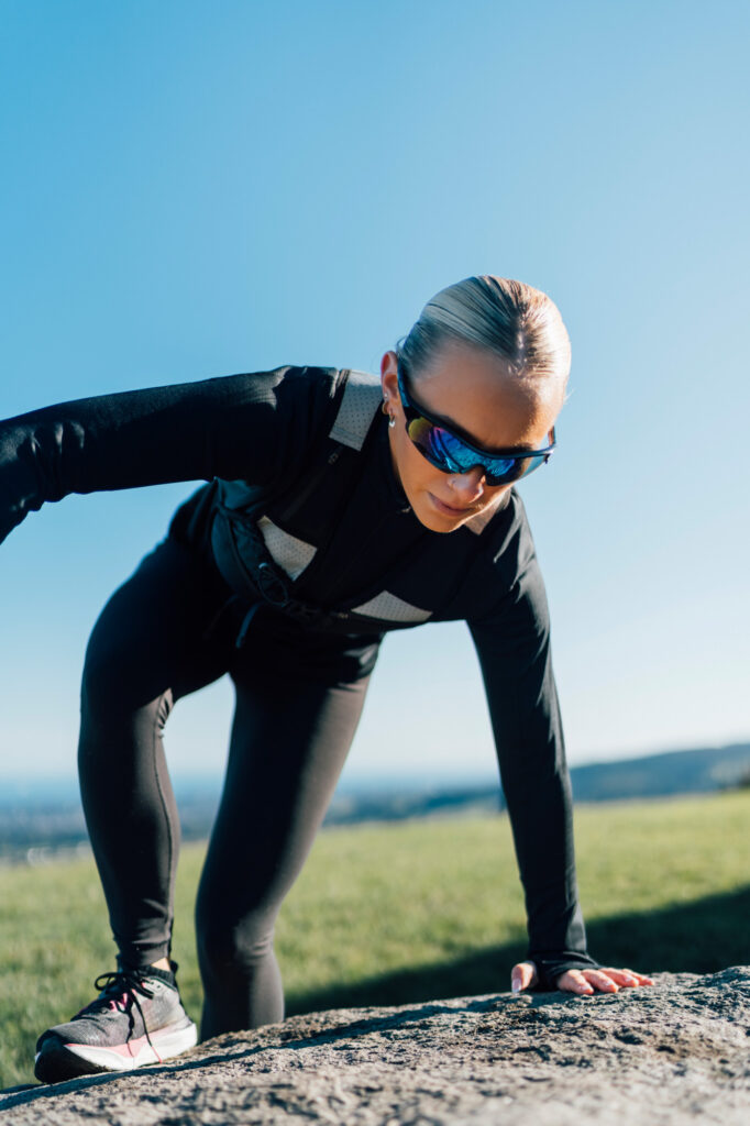 A trail runner crouches on a rocky edge, adjusting her stance in preparation for the next move. The open countryside and blue sky provide a scenic backdrop for outdoor adventure.