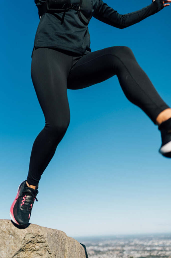 A trail runner leaps energetically on a hilltop, arms raised in motion, with a vibrant blue sky and scenic countryside in the background. The image highlights freedom and strength