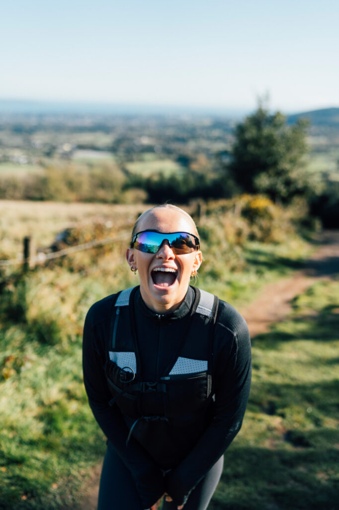 A female trail runner bursts into laughter while standing in the Irish countryside, surrounded by hills and greenery. The joyful moment highlights energy, freedom, and connection to nature.