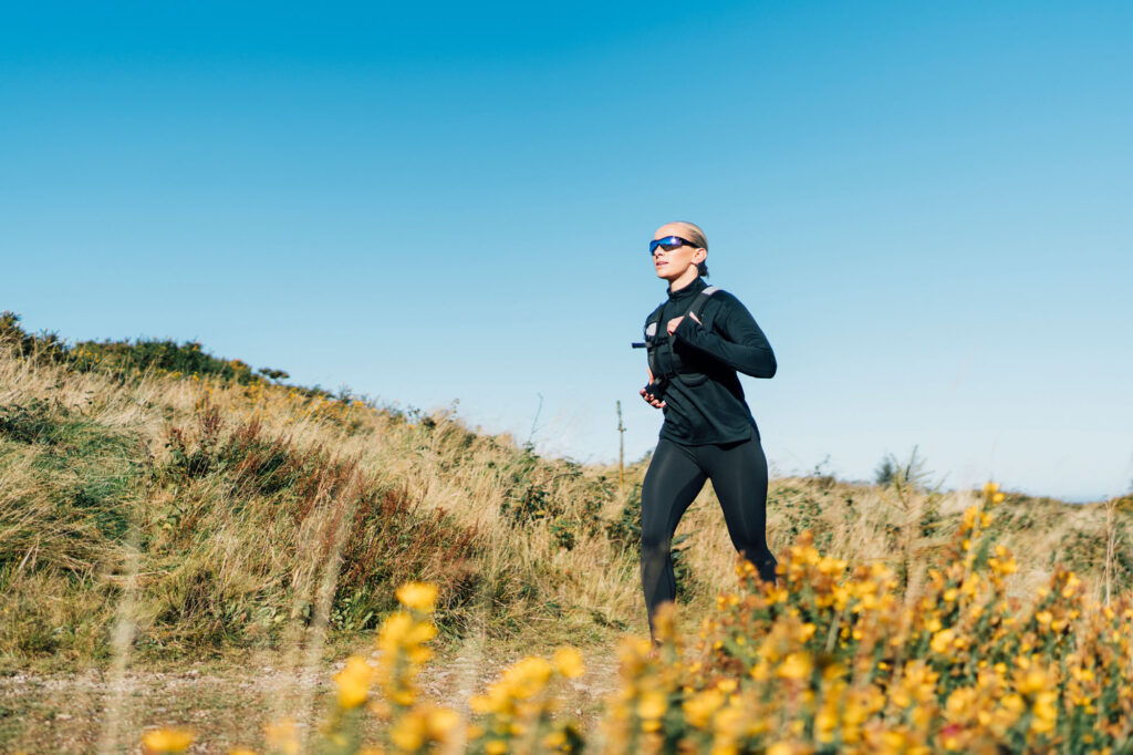 A trail runner strides confidently across a grassy field, surrounded by wildflowers and a bright sky. The image celebrates outdoor energy and freedom.